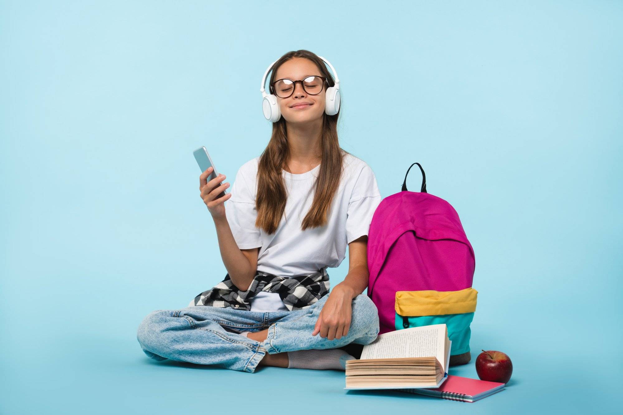 Cheerful caucasian schoolgirl teenager pupil student listening to the music song singer playlist in headphones while preparing going back to school isolated in blue background